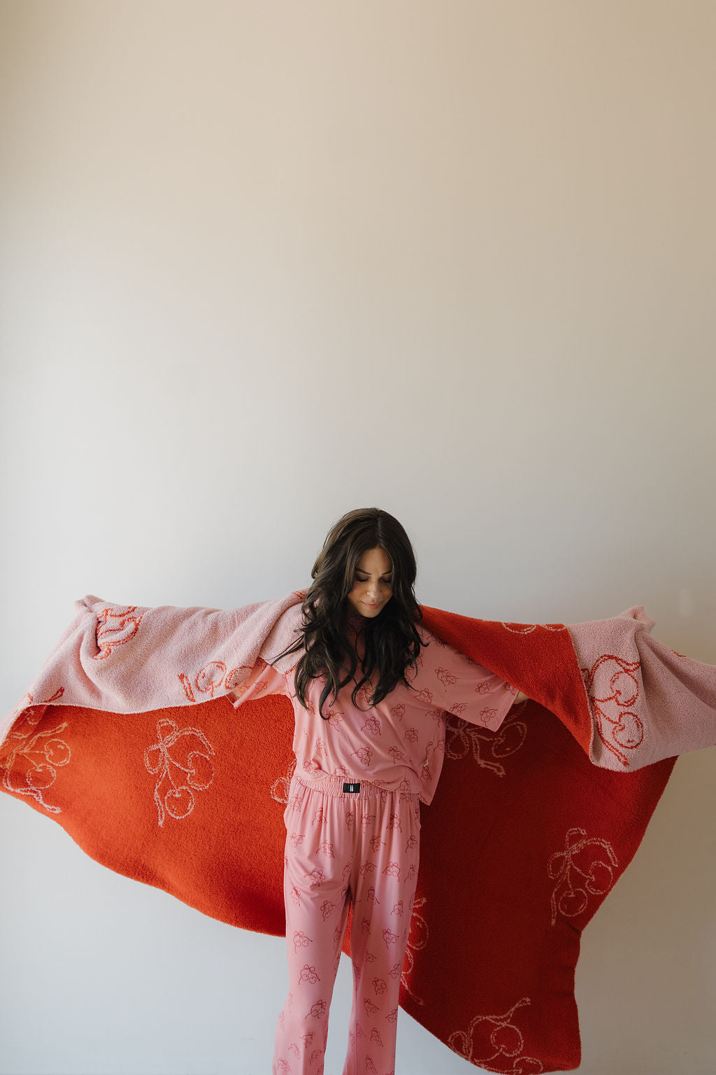 A woman wearing pink pajamas with red prints stands with her arms outstretched, draping a red and pink blanket behind her, against a plain light-colored wall.
