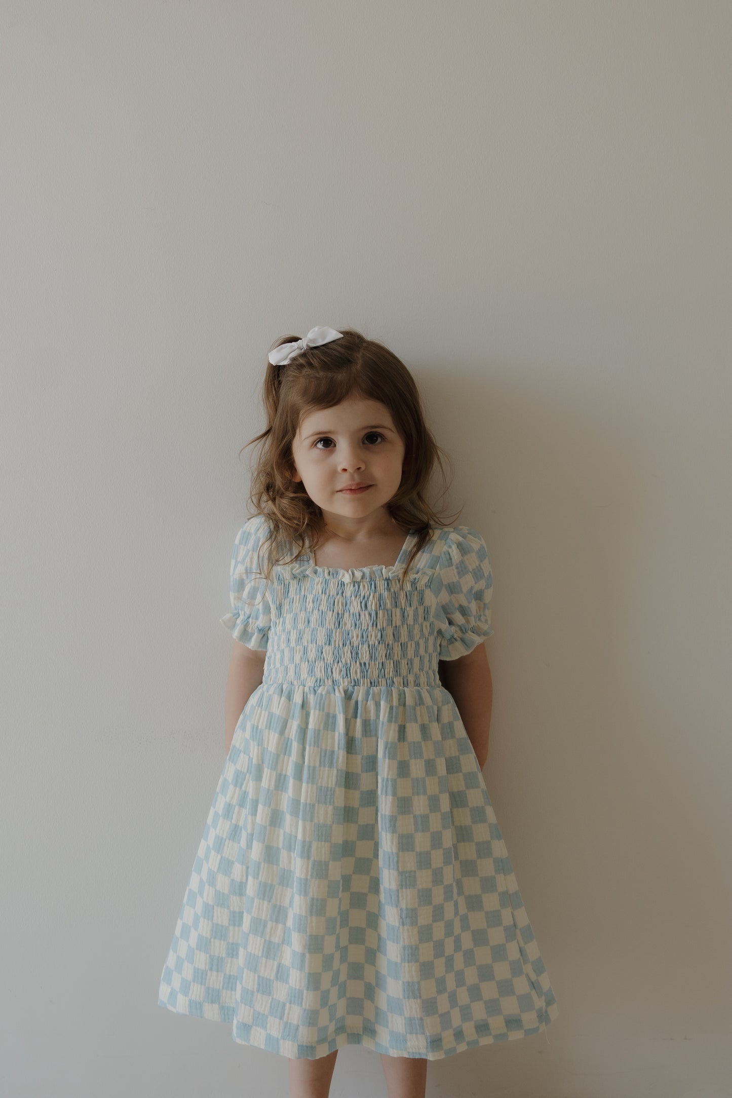 A young girl with light brown hair and a white bow wears the forever french baby Child Muslin Dress | Morning Check, featuring a blue and white checkered pattern, and stands against a plain light wall, smiling slightly at the camera.