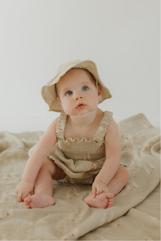 A baby in the forever french baby Sleeveless Muslin Romper & Hat | Silly Goose sits on a beige blanket, looking up with curiosity against a plain light background.