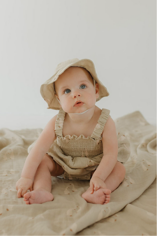 A baby in the forever french baby Sleeveless Muslin Romper & Hat | Silly Goose sits on a beige blanket, looking up with curiosity against a plain light background.