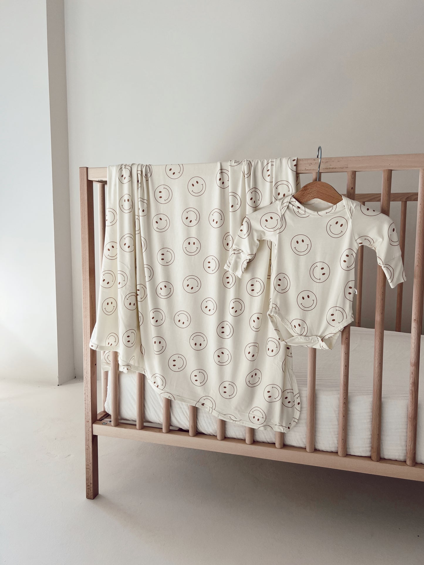 A minimalist nursery scene showcases a wooden crib with a white mattress. Draped over the crib is the "Bamboo Swaddle | Just Smile" from forever French baby, accompanied by a coordinating baby outfit. Both items offer year-round comfort and are embellished with simple, smiling faces. The walls and floor are adorned in light, neutral tones.