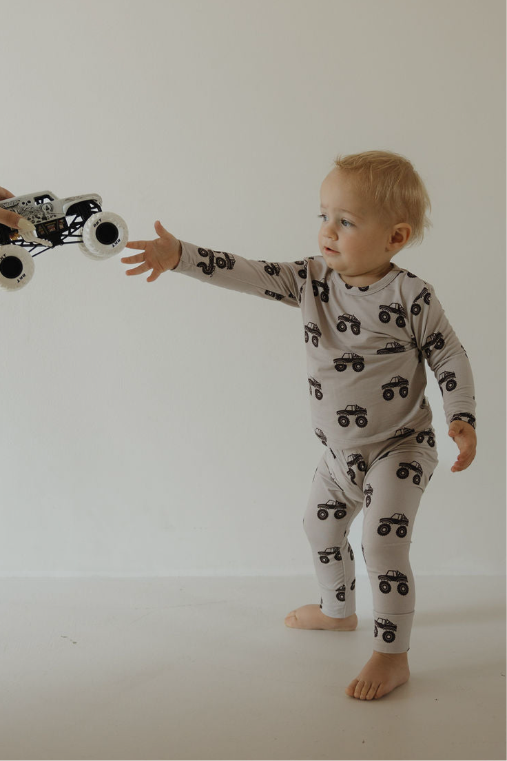 A toddler in the forever french baby Bamboo Footless Sleepy Time Set | Monster Truck reaches out for a toy monster truck being handed to them against a plain, light background.