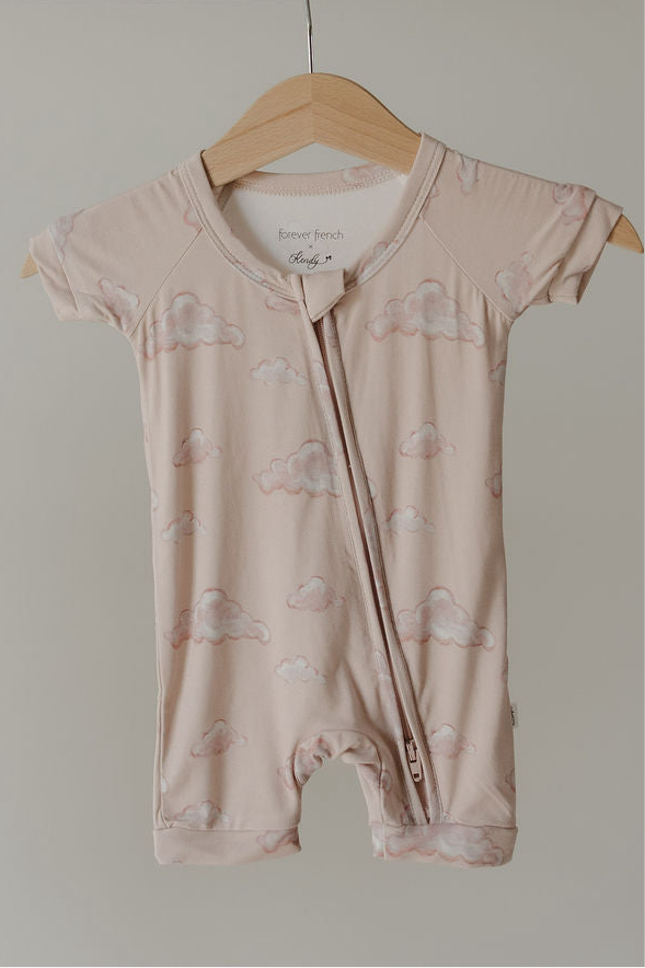 The Kendy 3 Bamboo Zip Shortie | Ruby is a short-sleeved, light pink baby romper with a cloud pattern, made from premium bamboo pajamas material and featuring a diagonal zipper from collar to left leg. Shown on a wooden hanger.