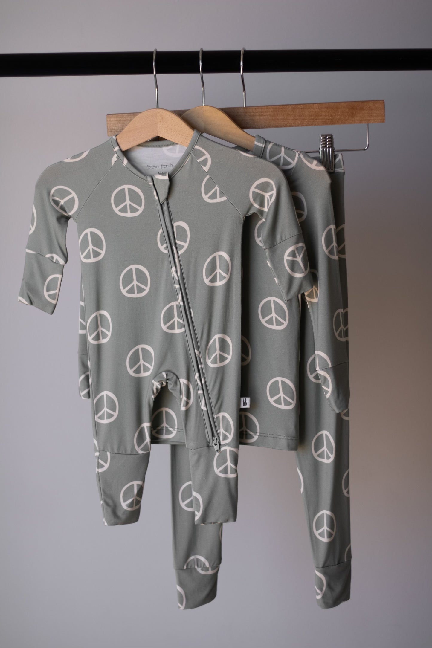 Two green Bamboo Zip Pajamas | Peace by forever french baby, featuring white peace signs, hang on wooden hangers from a black rack. Made from breathable baby sleepwear fabric—one is a zip-up onesie, the other a two-piece set.