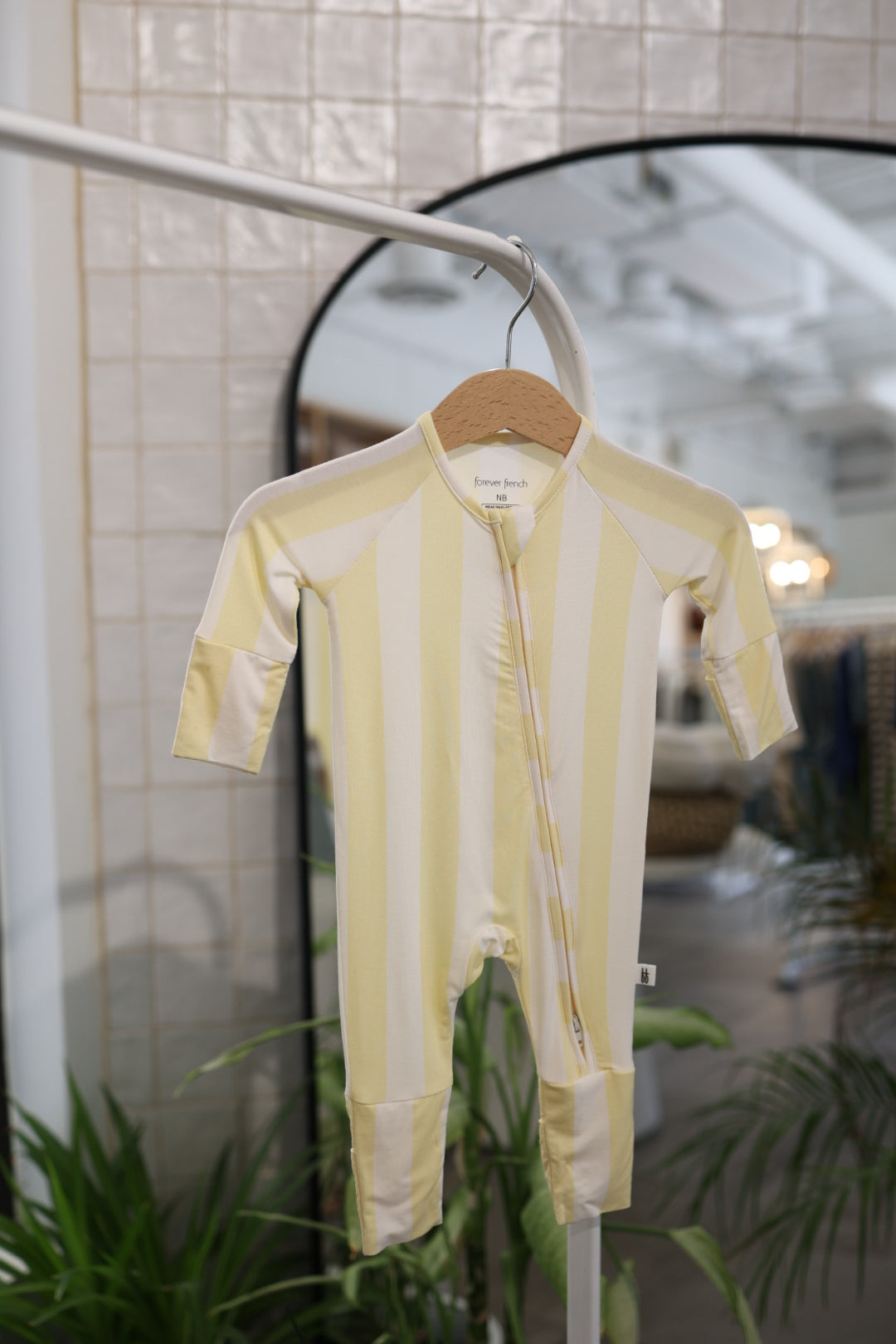 The Bamboo Zip Pajamas | Butter by forever french baby—a yellow and white striped, breathable baby onesie with a zipper—hangs on a wooden hanger in front of a mirror, white tiled wall, and green indoor plants.
