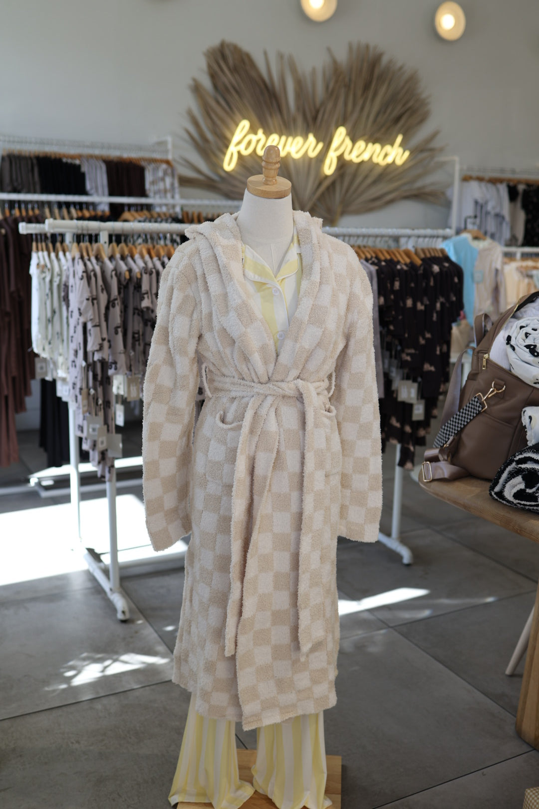 A mannequin wears the forever french baby Adult Robe in Cream Checkerboard and yellow-striped pajamas. Clothing racks and a neon "forever french" sign are visible in the background.