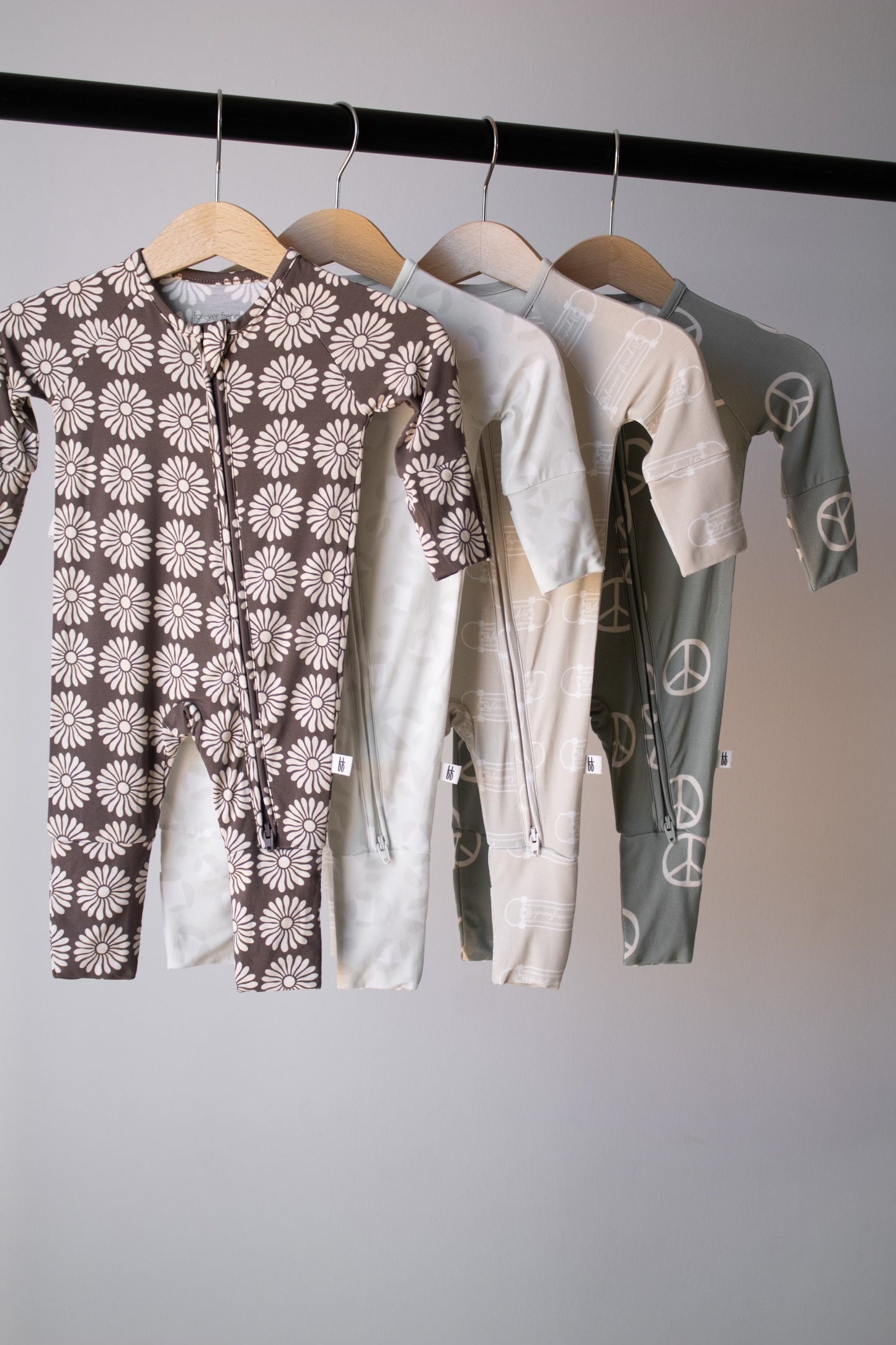 Four Bamboo Zip Pajamas | Retro Floral by forever french baby, featuring daisy and peace sign patterns, hang on wooden hangers from a black rod against a plain light gray background.