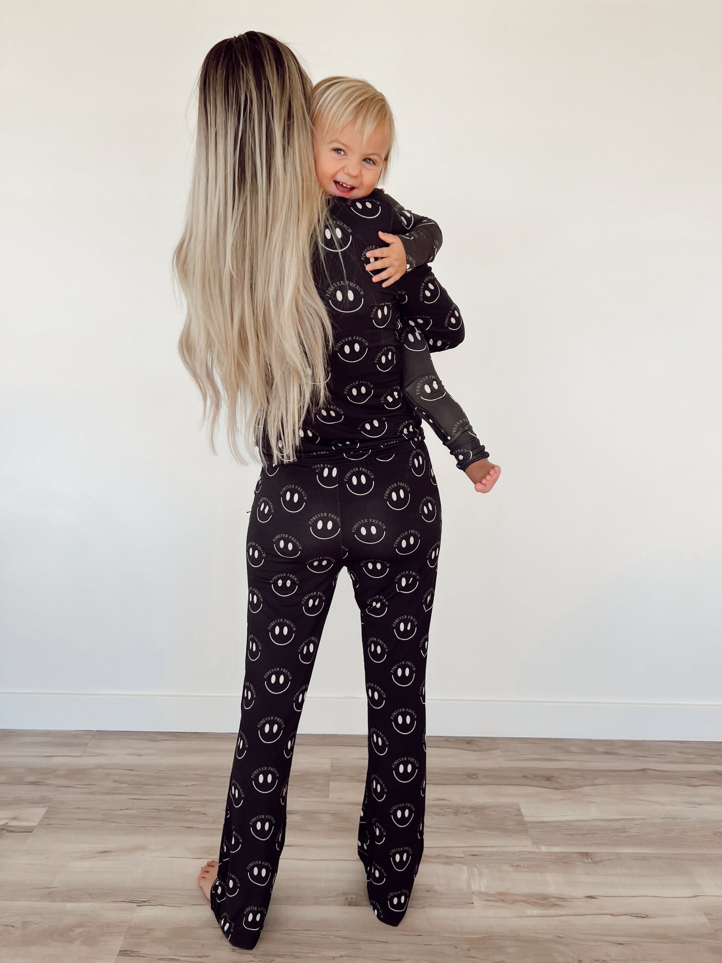 A woman with long blonde hair, seen from the back, holds a smiling toddler. Both are wearing matching pajamas from forever french baby, specifically the Women's Bamboo Pajamas in Charcoal & White ff Smile. Known for their year-round comfort and hypo-allergenic properties, they stand on a wooden floor against a plain white wall. The toddler looks over the woman's shoulder and smiles at the camera.