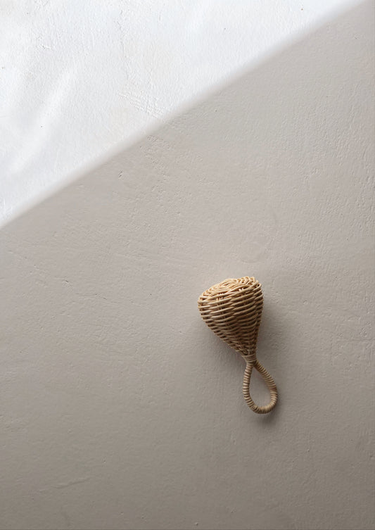 A small, textured, cone-shaped hanging ornament made of tightly coiled natural fibers resembles the Rattan Baby Rattle by forever french baby. It is mounted on a plain light-colored wall that is diagonally divided by a shadow, creating a contrast between two shades of the same color.