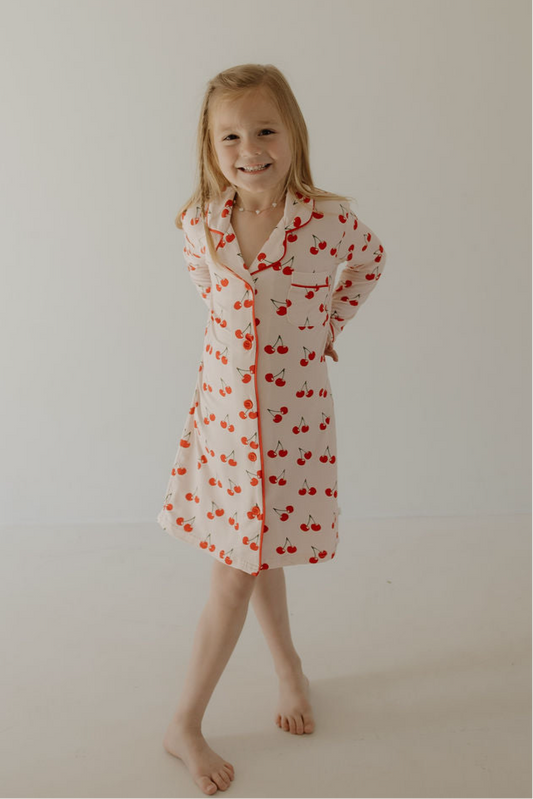 A smiling young girl with long blonde hair stands barefoot against a plain background, wearing the forever french baby Toddler Bamboo Sleeping Dress | Cherry Orchard, made from soft hypo-allergenic fabric. She looks relaxed and happy with her hands behind her back.