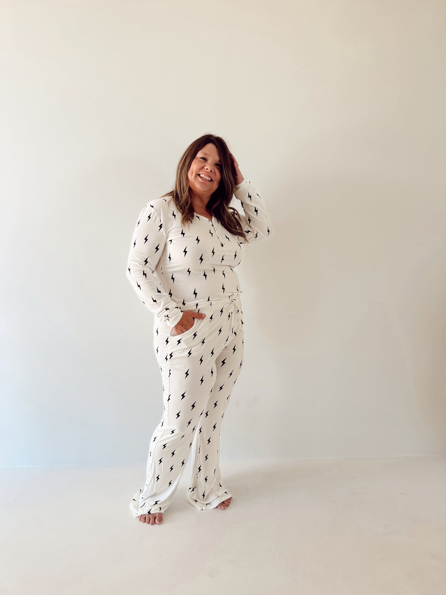 A woman stands against a plain white background, smiling with one hand touching her hair. She is wearing the Women's Bamboo Pajamas by forever french baby, featuring a simple, small black and white lightning bolt print throughout. Her other hand is in her pocket, and she is barefoot.