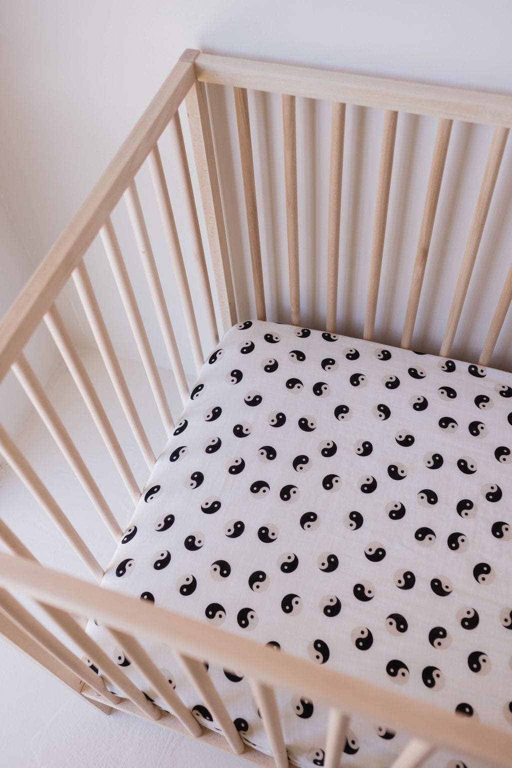 A wooden crib with vertical slats is shown with a forever french baby Muslin Crib Sheet | Black & Ivory Yin Yang. The setting is minimal and bright, suggesting a calm and serene environment.