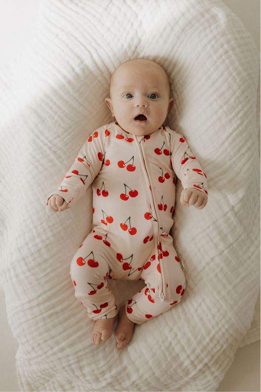 A baby lies on a soft white blanket, wearing forever french baby's Bamboo Zip Pajamas in the Cherry Orchard print—cream-colored with a red cherry pattern and made from breathable fabric—looking up with bright, alert blue eyes.