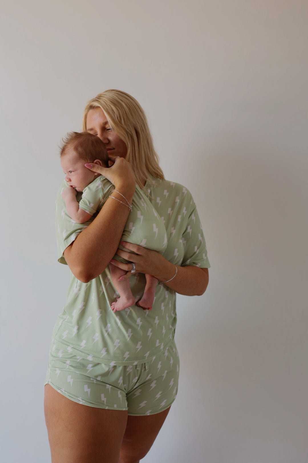 A woman wearing the forever french baby Women's Bamboo Short Set | Flash holds a baby in matching green lightning bolt bamboo outfits against a light background, both enjoying breathable comfort and affectionate moments together.