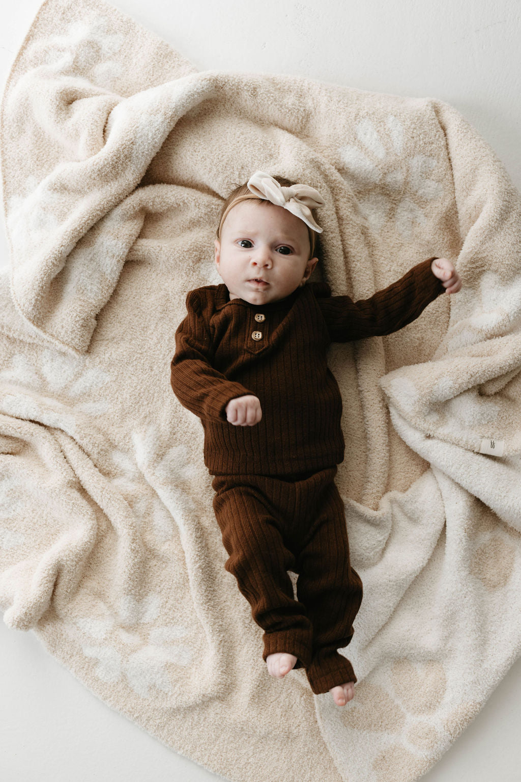 A baby wearing a brown knitted outfit and a beige headband with a bow lies on the Plush Blanket | Lazy Daisy by forever french baby, which has a velvety-rich texture. The baby is stretching one arm and looking towards the camera.