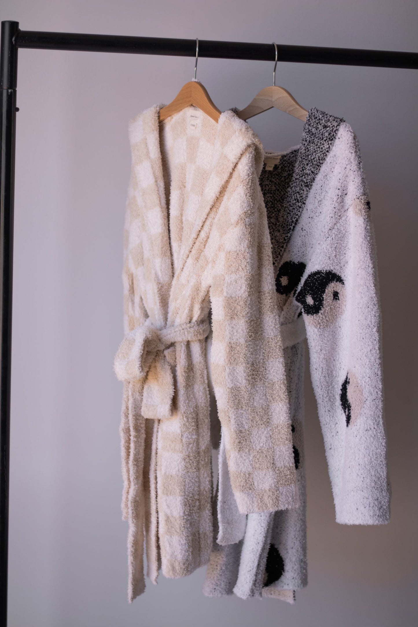 Two forever french baby bathrobes hang on a black rack: the Adult Robe | Cream Checkerboard in cream and white checks, and a white microfiber robe with a black yin-yang motif. Both are displayed on wooden hangers.