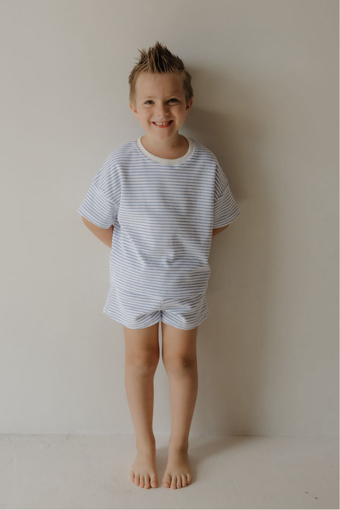 A smiling boy with short, spiky hair stands barefoot against a plain wall, wearing the forever french baby Child Terry Short Set | Ocean Stripe, capturing the joy of simple family moments.