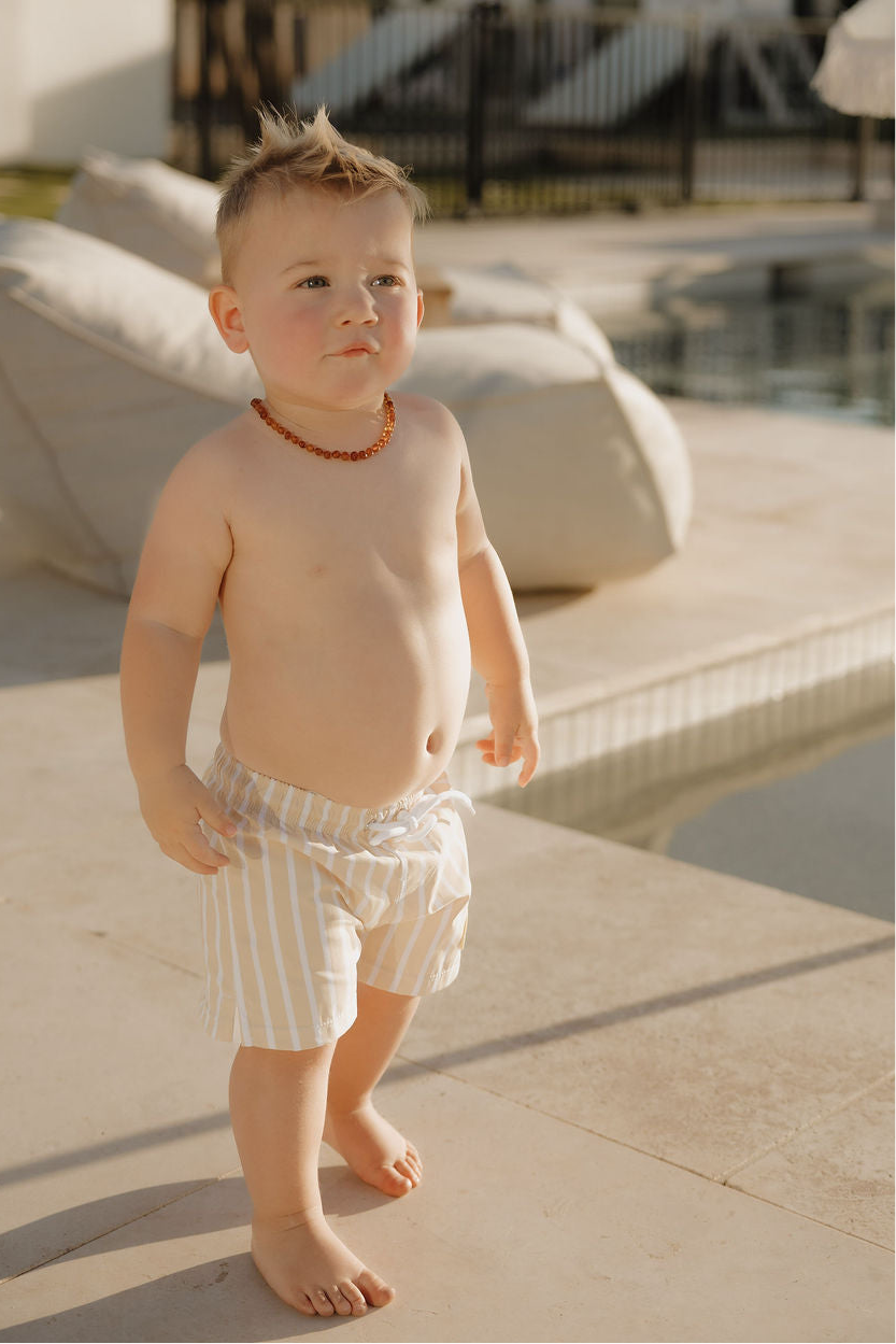 A young child with short light hair, barefoot and wearing lolo webb 2’s Child Board Short | Sun Rays and an amber necklace, stands by a pool on a sunny day.