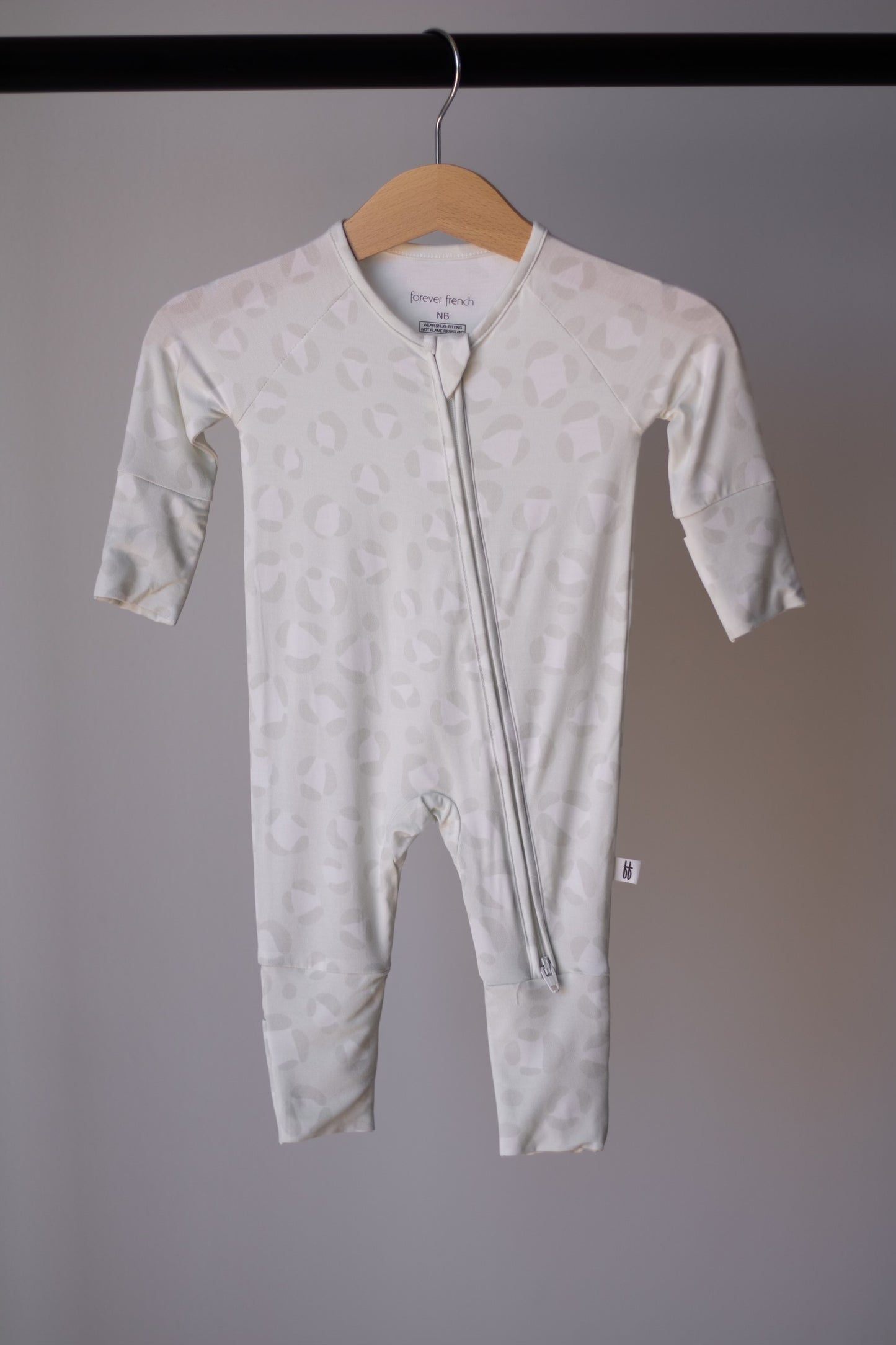The Bamboo Zip Pajamas | Meow by forever french baby is a light-colored, long-sleeve baby onesie with a subtle pattern and diagonal zipper, made from breathable fabric and shown on a wooden hanger against a plain background.