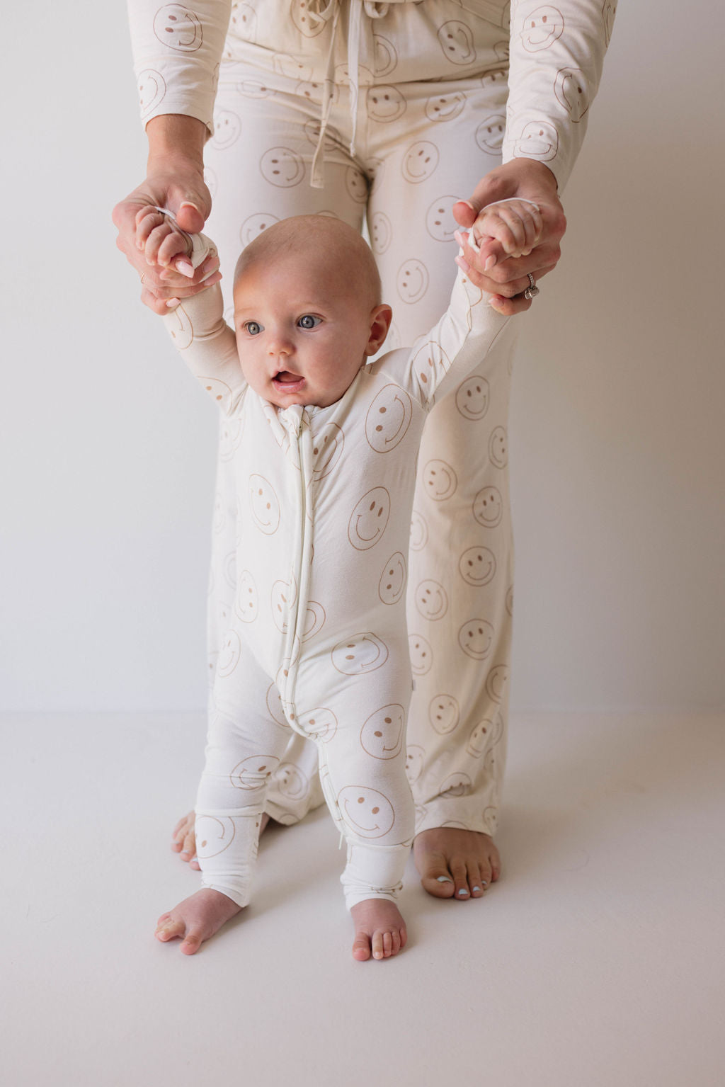 A baby dressed in the "Just Smile" hypo-allergenic white onesie with smiley face prints from forever french baby holds onto an adult's hands while trying to stand. The adult, also wearing matching "Just Smile" pajamas crafted from premium bamboo by forever french baby, supports the baby from behind. Both are barefoot on a white floor.