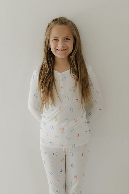 A young girl with long light brown hair wears Forever French Baby's Bamboo Two Piece Pajamas, featuring colorful numbers for autism awareness, and smiles at the camera with her hands behind her back against a white wall.