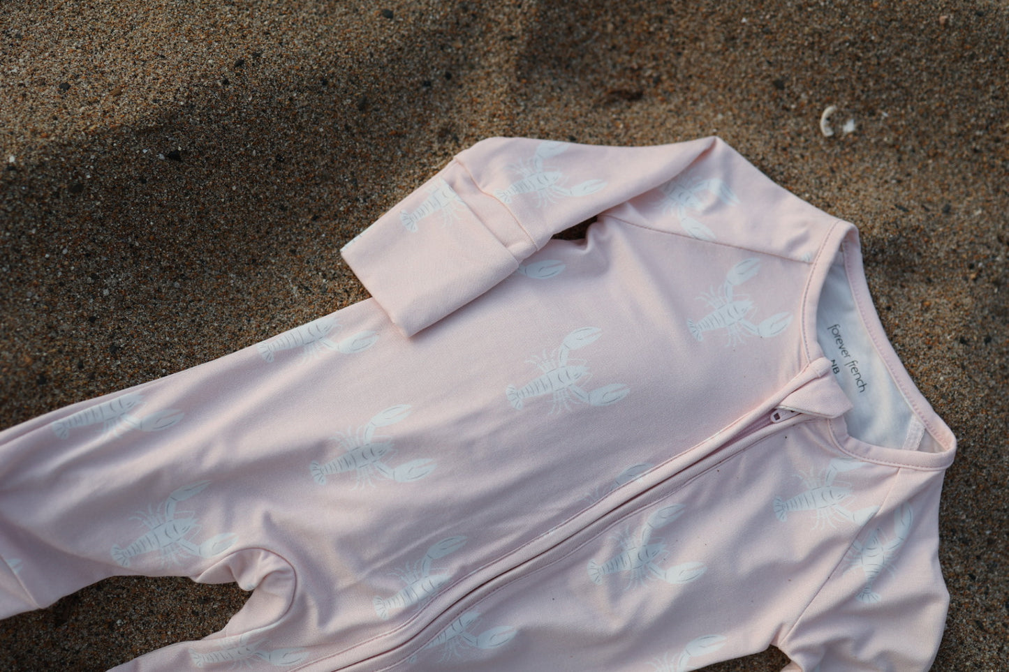 A pale pink Bamboo Zip Pajamas | Lobster by forever french baby, made from hypoallergenic fabric and featuring a white lobster print, lies partially buried on sandy ground.