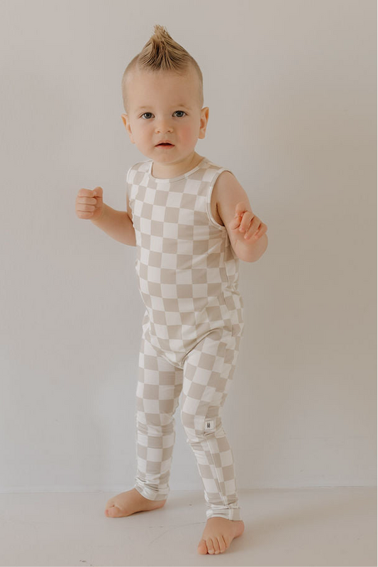 A young toddler with short, light hair in a mohawk stands barefoot on a light floor, wearing the forever french baby Harem Bamboo Romper | Checkerboard—a sleeveless, beige-and-white checkered piece made from breathable bamboo fabric.