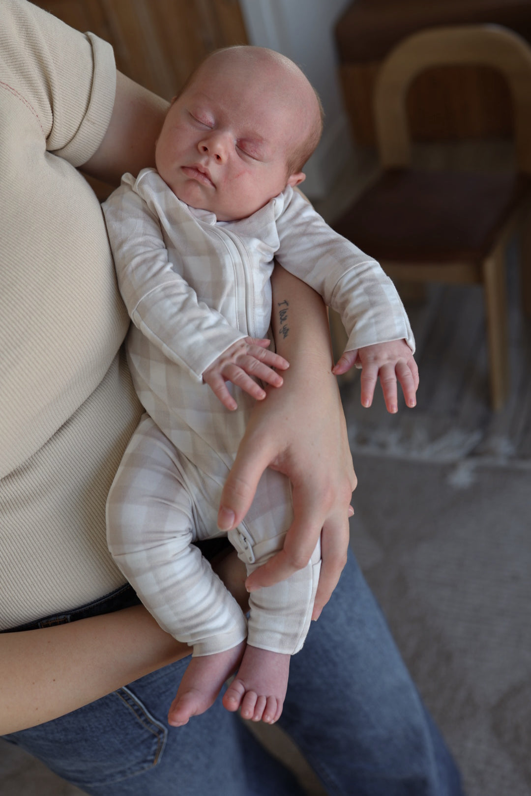 A baby, cozy in forever french baby's Bamboo Zip Pajamas | Gingham, sleeps peacefully while cradled by an adult dressed in a beige shirt and blue jeans. A wooden chair and rug are partially visible in the background.