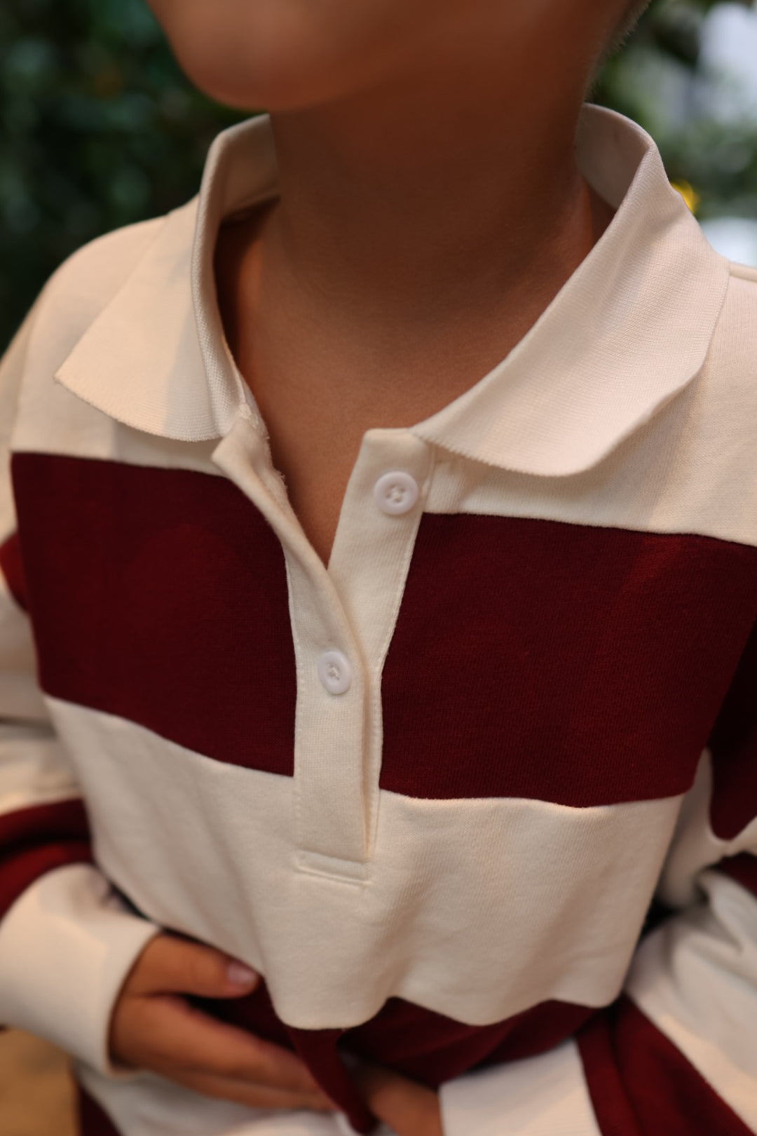 A child wearing a Child Rugby Sweatshirt | Deep Cranberry, a 100% cotton sweatshirt with wide, horizontal cranberry and cream stripes and a white collar with two buttons.
