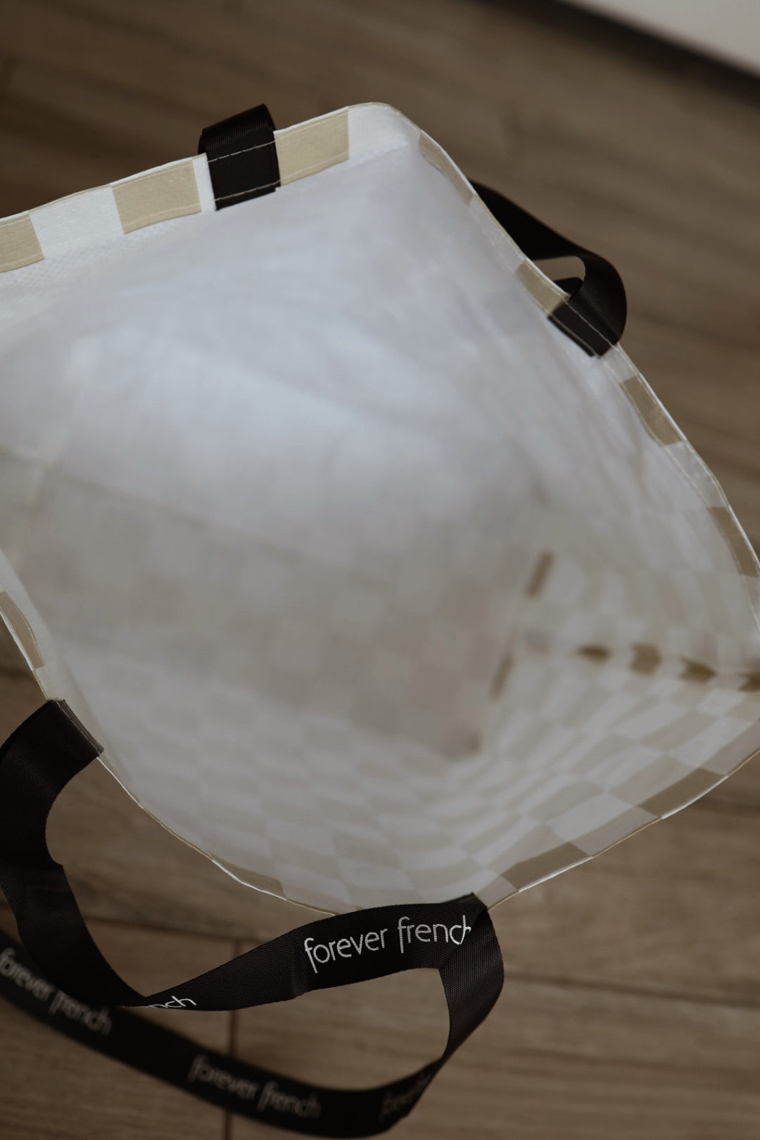 A top-down view of the ff Warehouse Sale Tote by forever french baby. The white checkered bag has black handles with "forever french" in white text, and rests on a wooden floor.