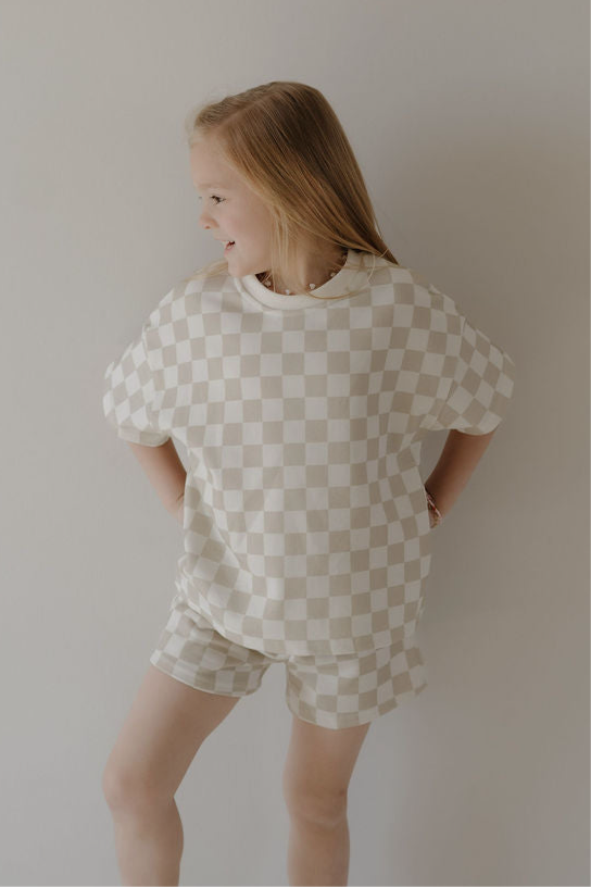 A young girl with long blonde hair smiles and looks to the side while wearing the forever french baby French Terry Short Set | Checkerboard, standing against a plain light-colored background.