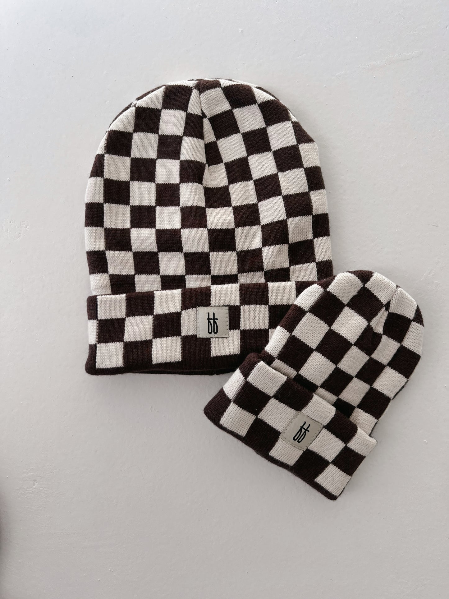 Two Coffee & Cream Checkerboard knit beanies from Forever French Baby are displayed against a white background. The larger beanie has a folded brim showcasing its classic ribbed design, while the smaller one is more narrow. Both feature a black and white checkered pattern and a small tag with the letters "bb" on the front.