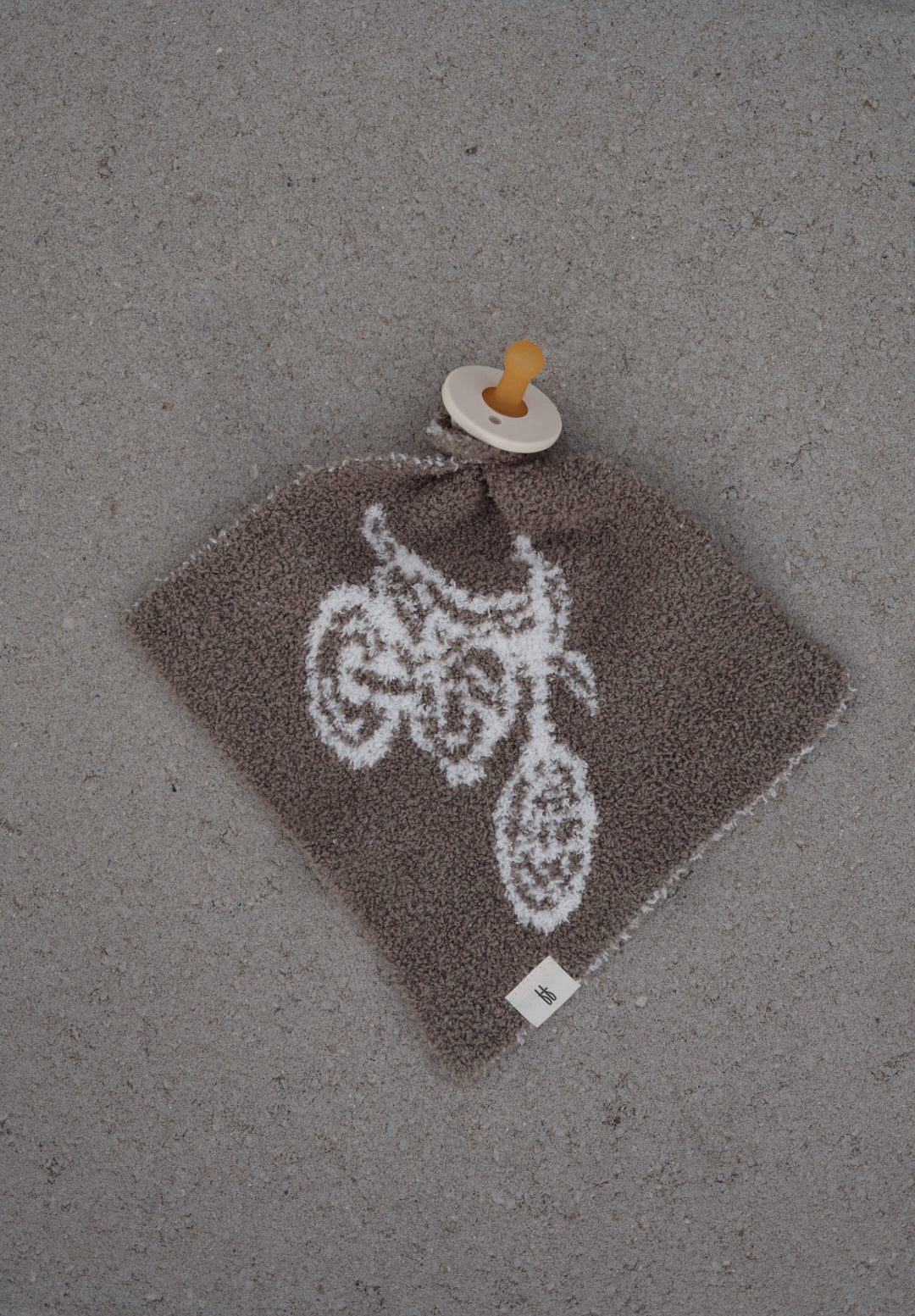 A Lovey | Dirt Bike by forever french baby, featuring a brown textured fabric with a white dirt bike design and attached to a yellow pacifier, rests on a gray surface.