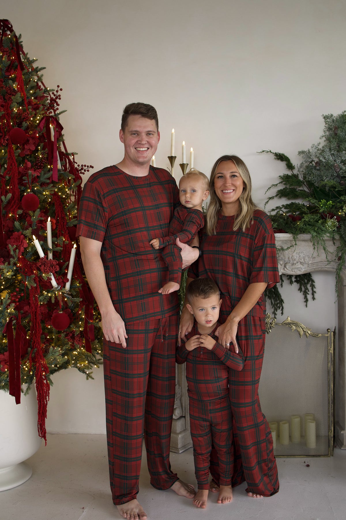 A family of four stands together in matching red and green plaid Men’s Short Sleeve Pajamas | Christmas Plaid by forever french baby in front of a decorated Christmas tree.