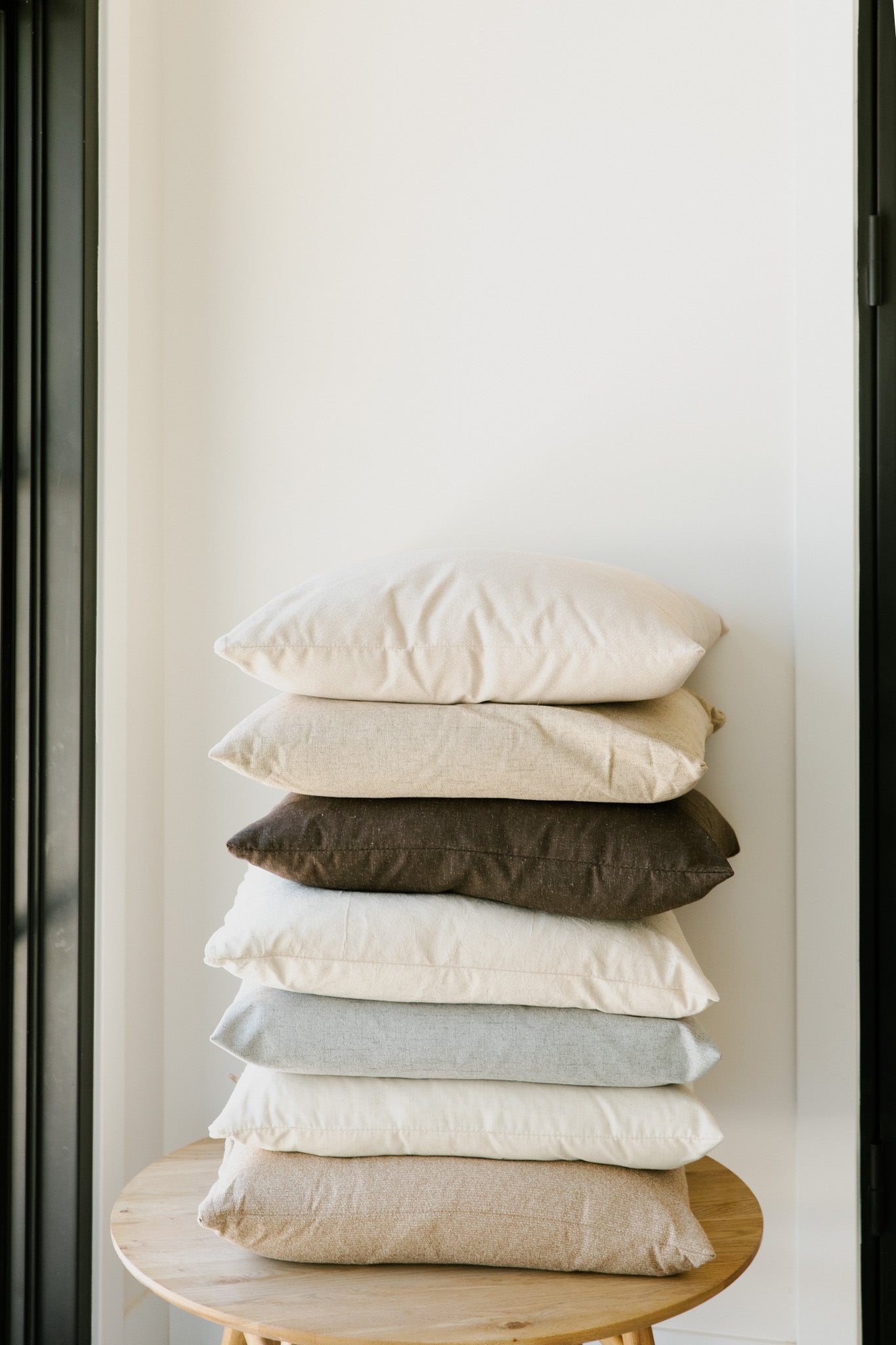 A stack of six differently colored Limestone | French Pillows from forever french home is placed on a round wooden stool in front of a plain white wall. The pillows, each with a feather down insert, are arranged neatly, with colors ranging from light beige at the top to dark brown in the middle and soft blue at the bottom.