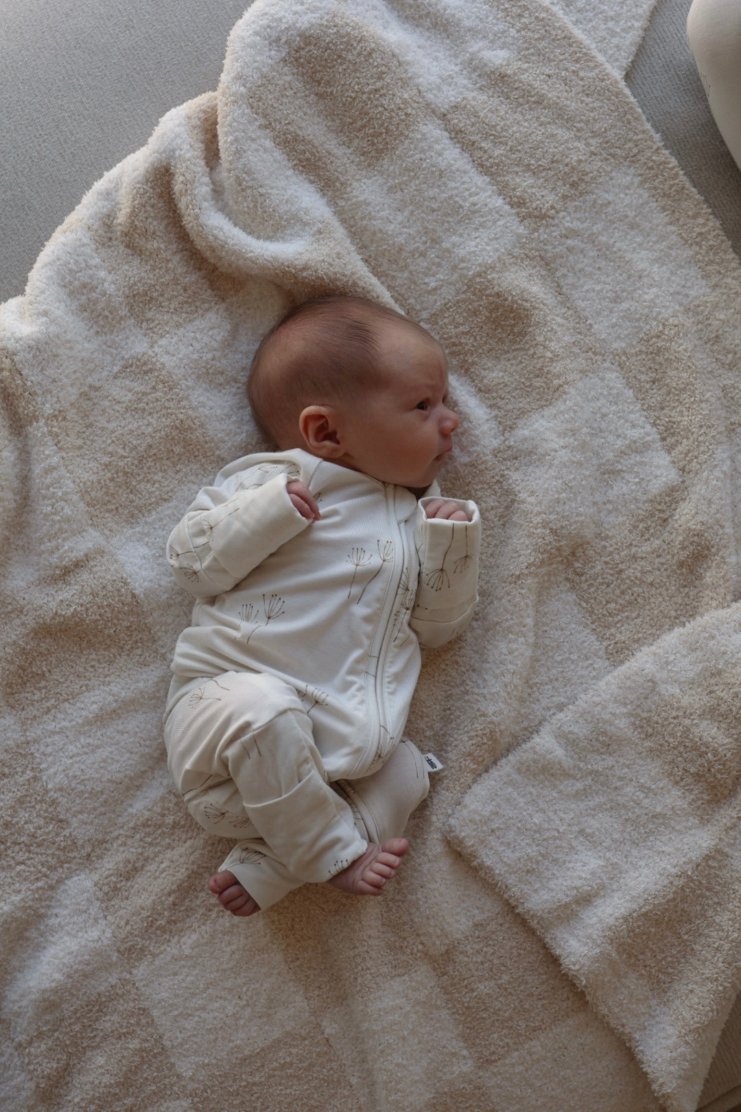 A baby wearing forever french baby's Bamboo Zip Pajamas in Dandelion lies on a textured cream-colored blanket, gazing to the side with hands near their face and feet curled up.