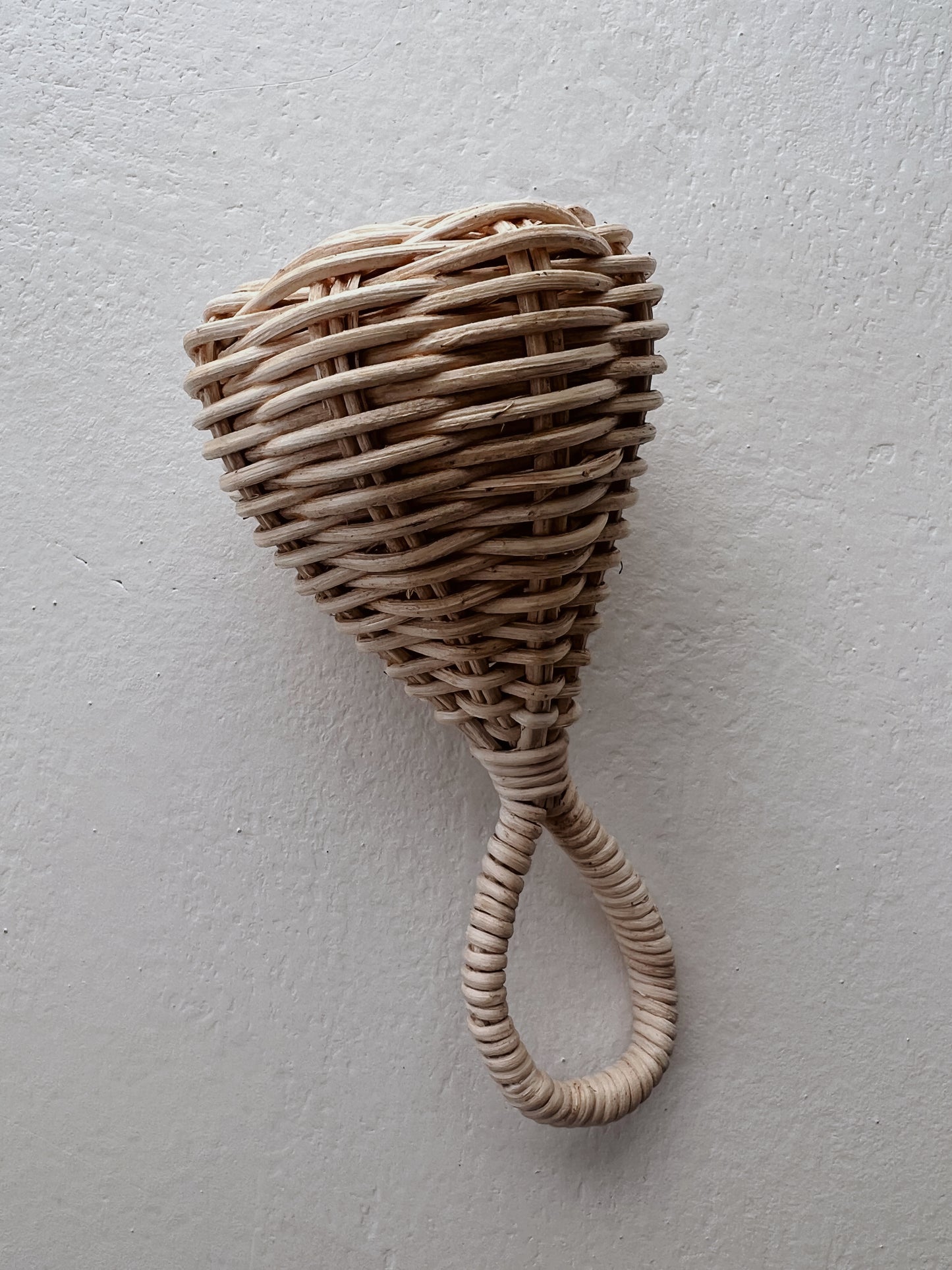 A Rattan | Baby Rattle from forever french baby hangs against a plain, light-colored wall. This handmade baby rattle features a teardrop-shaped head and a circular handle, both intricately crafted from natural rattan strands.