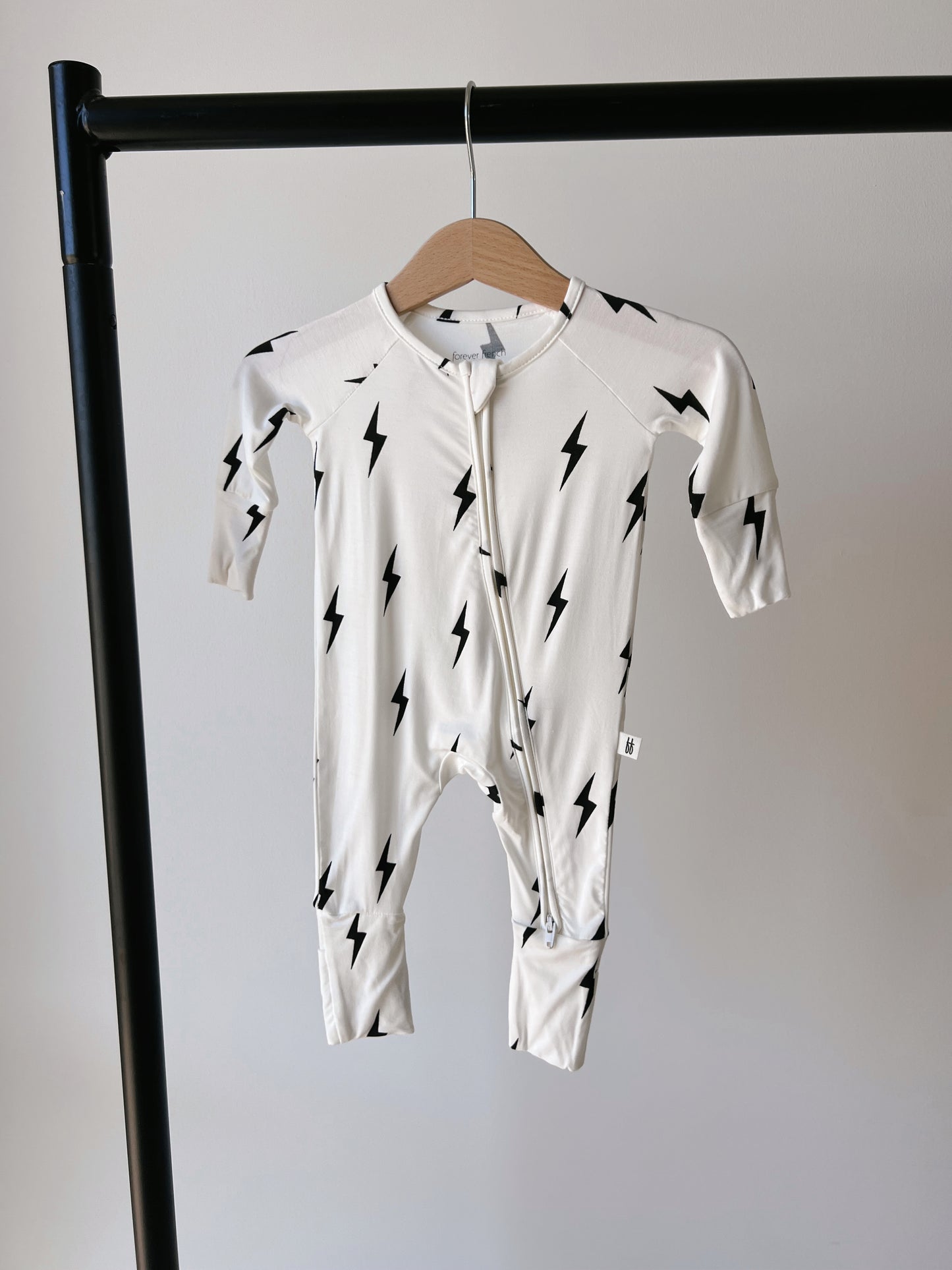 A cream-colored baby onesie with black lightning bolt patterns is hanging on a wooden hanger, displayed on a black clothing rack against a plain white background. This Forever French Baby Bamboo Zip Pajamas in White & Black Lightning Bolt features a front zipper closure and boasts their distinctive lightning bolt design.