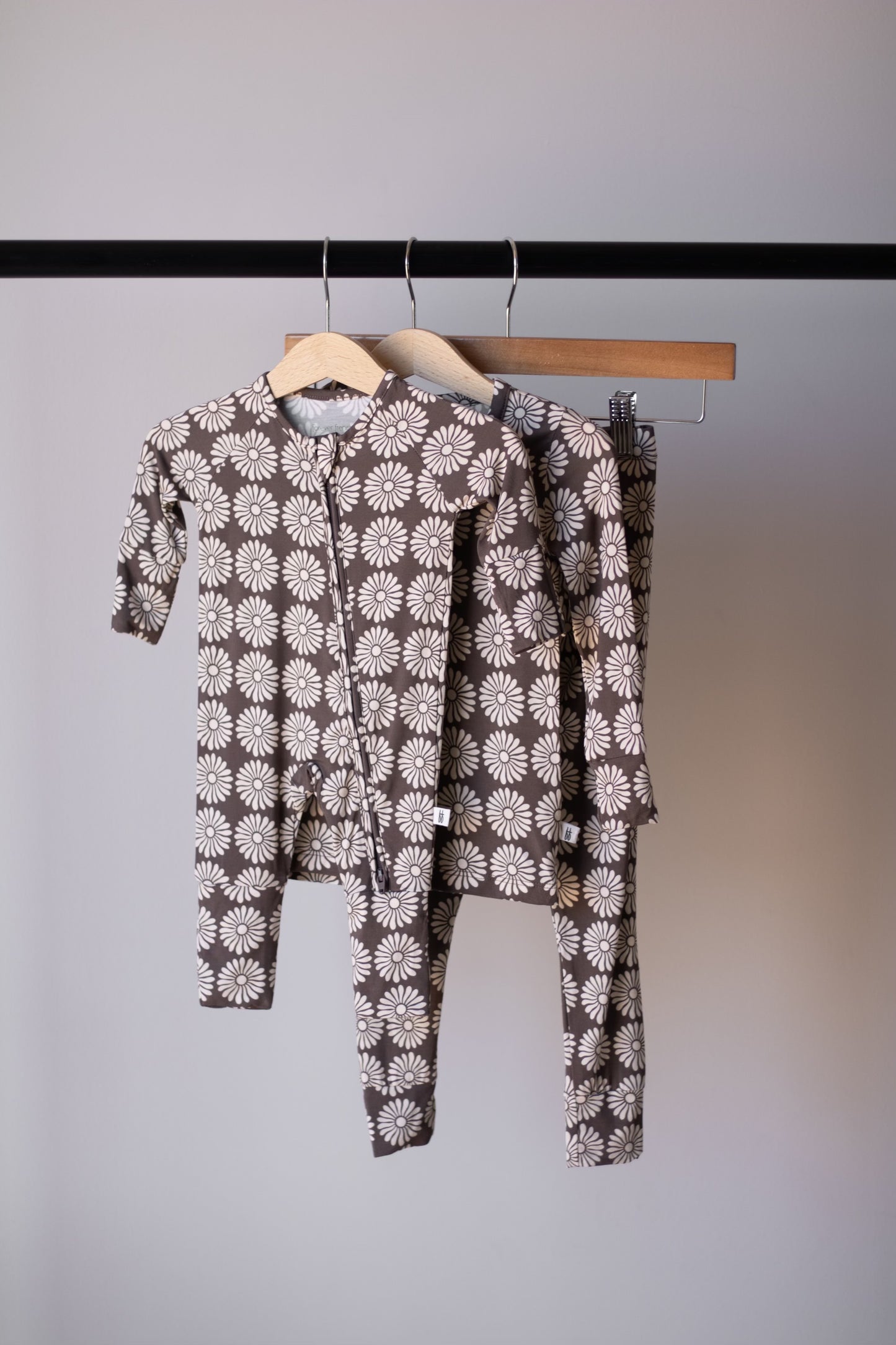 Two matching forever french baby Bamboo Zip Pajamas in Retro Floral, one adult and one child-sized, with white daisy patterns, hang on wooden hangers against a plain gray wall.
