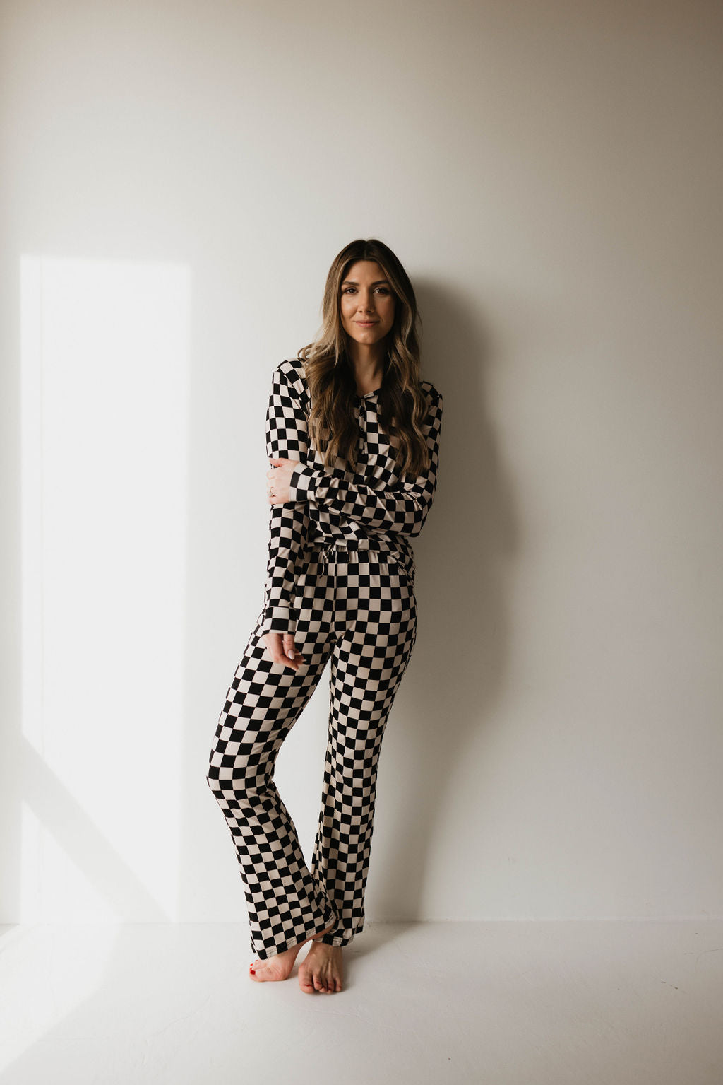 A woman with long brown hair is leaning against a white wall. She is barefoot and dressed in a matching black and white checkered long-sleeve top and pants set, identified as Women's Bamboo Pajamas | Black Checkerboard by forever french baby. Sunlight streams in from a window on the left side of the image, casting a soft shadow.