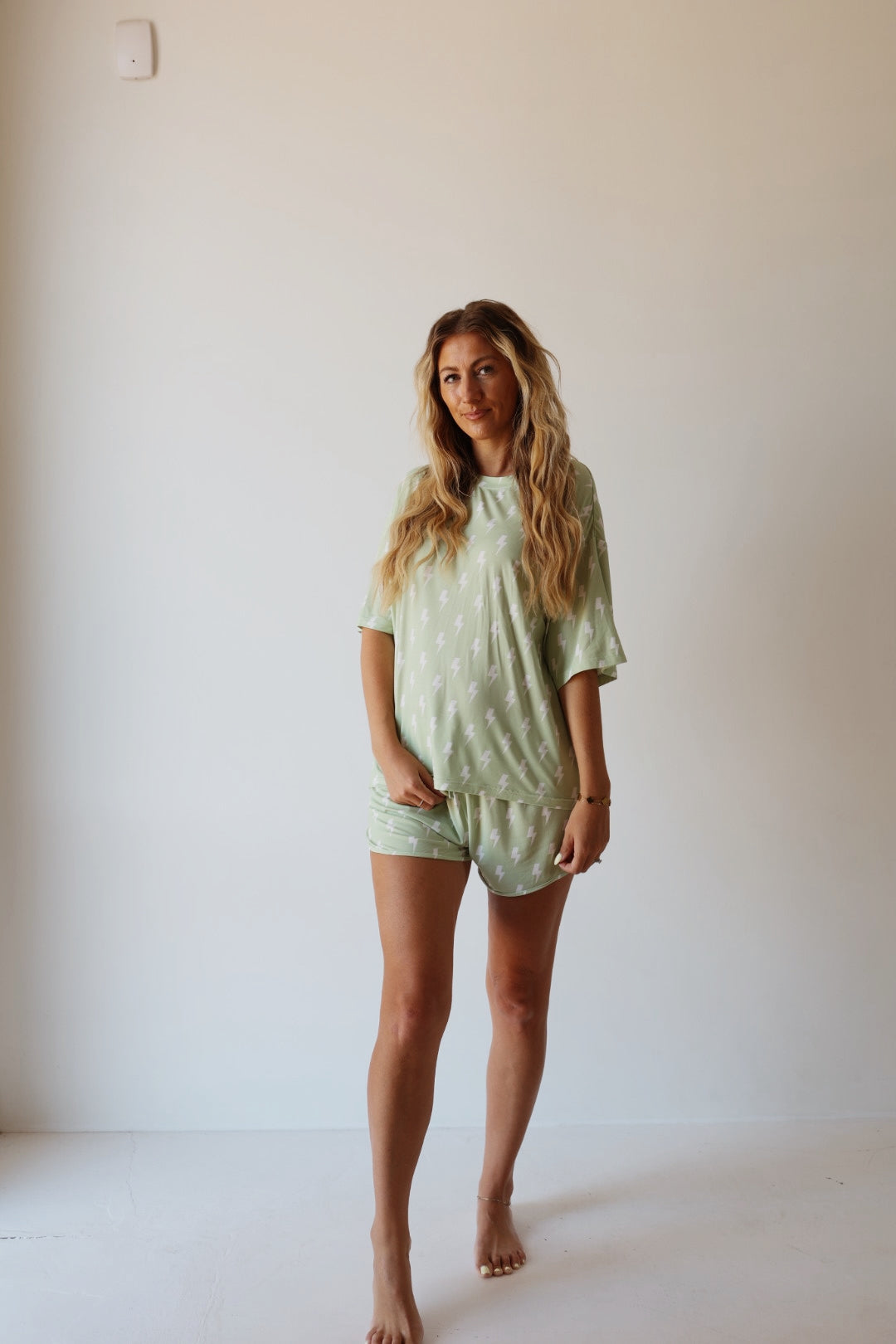 A woman with long, wavy blonde hair stands barefoot by a light wall, smiling softly at the camera. She wears the forever french baby Women's Bamboo Short Set | Flash—a light green, breathable pajama set with a subtle white pattern.