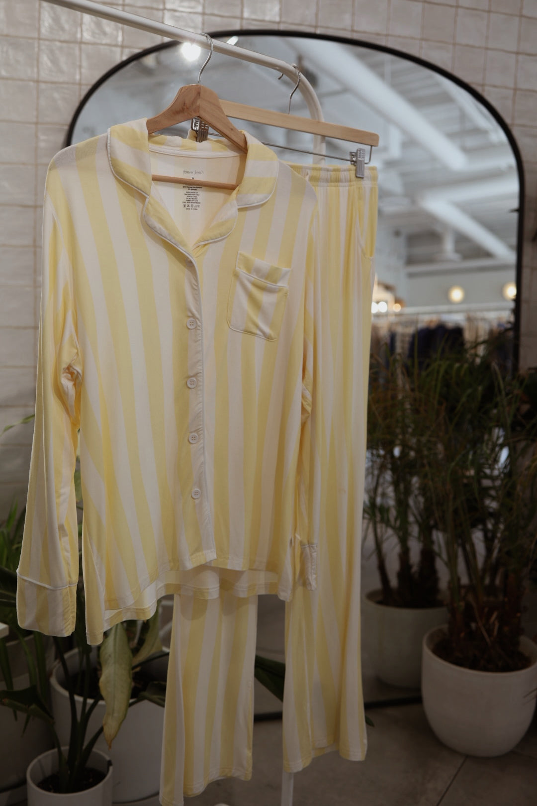 The Long Sleeve Women's Bamboo Pajama in Butter by forever french baby, with yellow and white stripes, a button-up top, and lounge pants, hangs on a wooden hanger before a mirror, amid potted plants and a tiled wall.