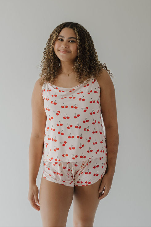 A woman with curly hair smiles, wearing the forever french baby Cami Women's Bamboo Set | Cherry Orchard—cooling bamboo pajamas with a cherry print—standing against a plain light-colored wall.
