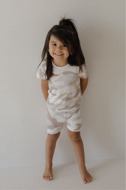 A young child with long dark hair stands barefoot against a light wall, smiling and wearing the forever french baby Bamboo Short Set | Camo 2.0—a matching light camo t-shirt and shorts made from breathable fabric for all-day comfort.
