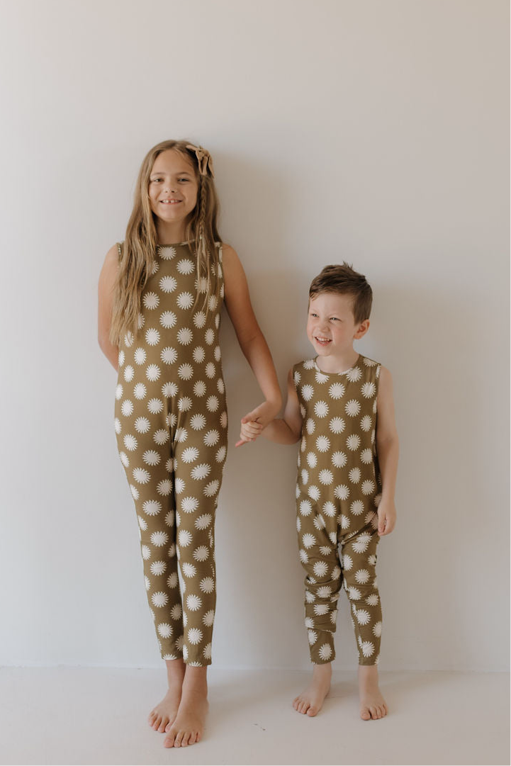 Two young children stand barefoot against a white wall, holding hands and smiling in matching forever french baby Harem Bamboo Rompers | Sol—daisy print outfits designed for comfort, playfulness, and breathable style.