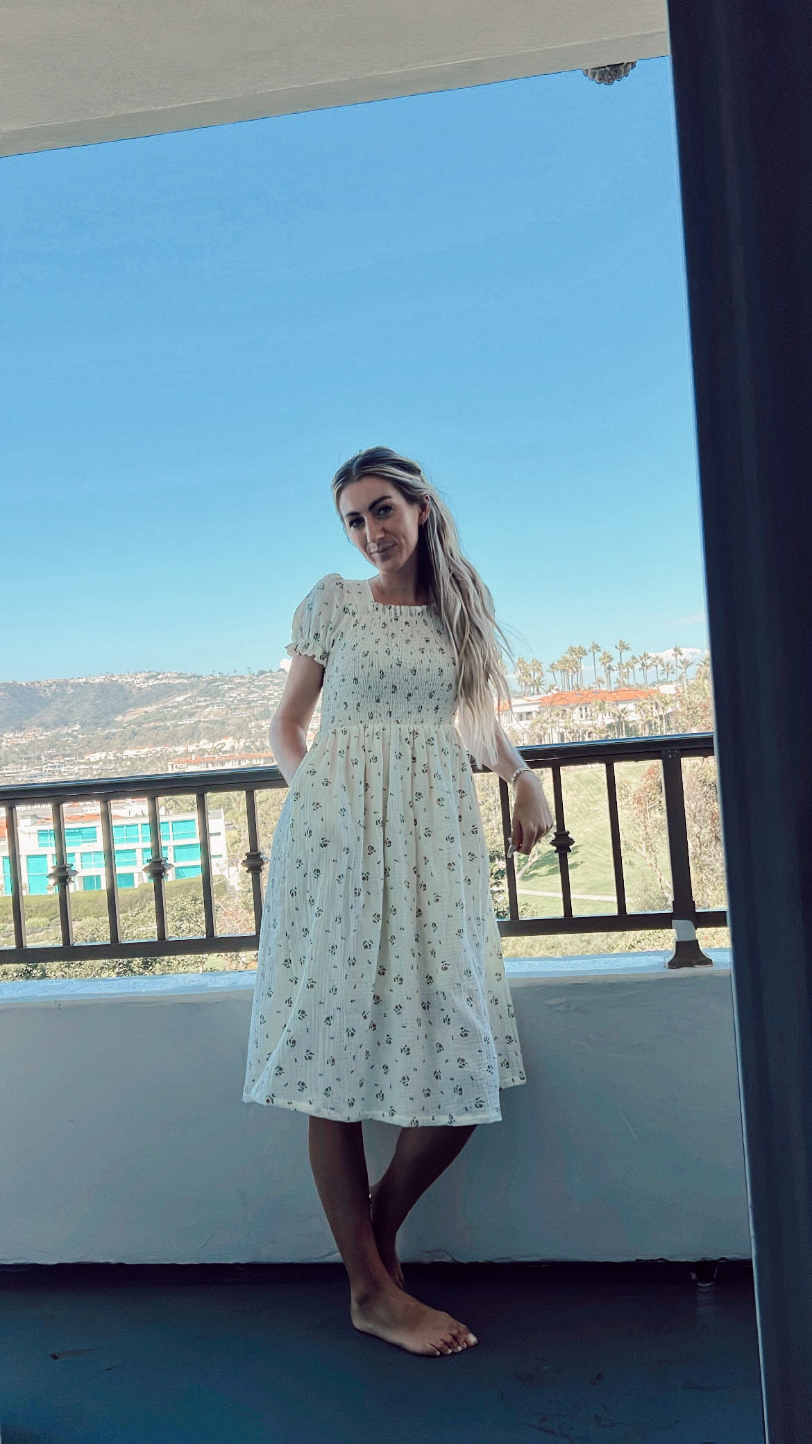 A woman with long hair stands on a balcony, barefoot and leaning against the railing, wearing a Garden Days ivory muslin dress adorned with floral patterns by forever french baby. In the background, buildings, trees, and a clear sky are visible.