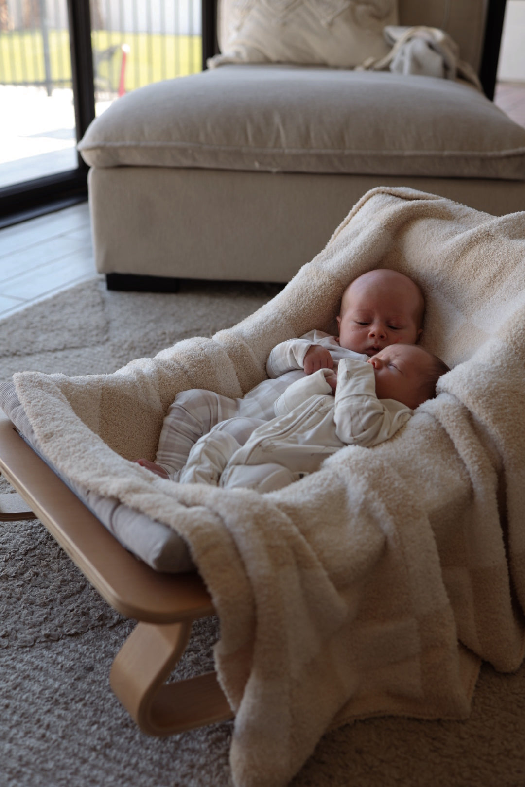Two sleeping babies rest side by side in a soft beige rocker, cozy in forever french baby Bamboo Zip Pajamas | Gingham. A plush chair and large windows with natural light fill the background.