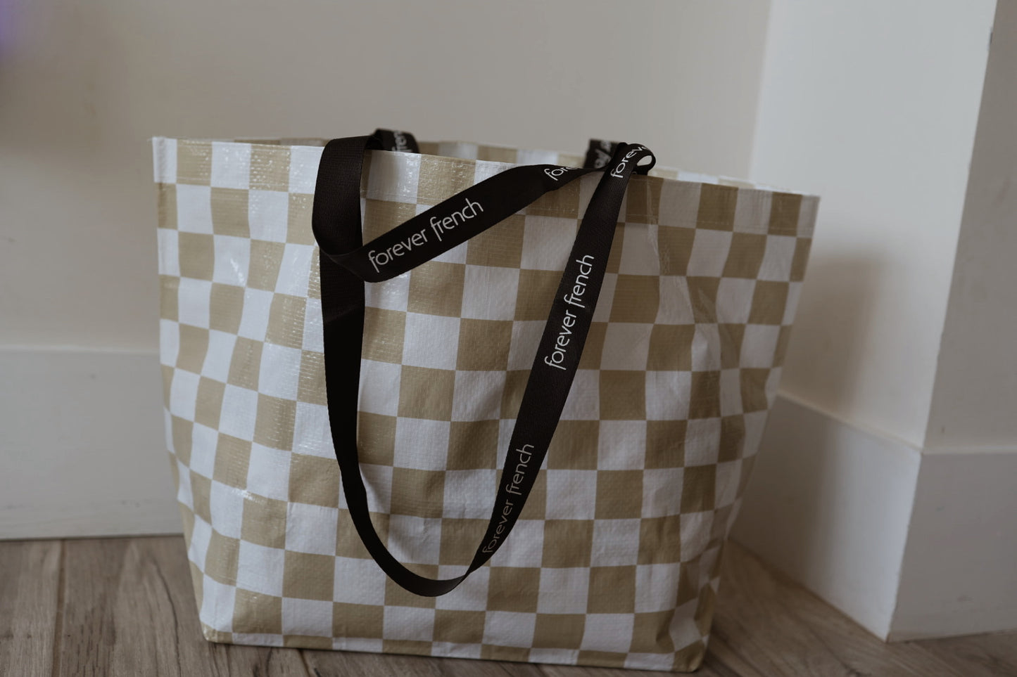The ff Warehouse Sale Tote by forever french baby features a beige and white checkered design with black handles, shown resting on a wooden floor near a white wall and corner baseboard.