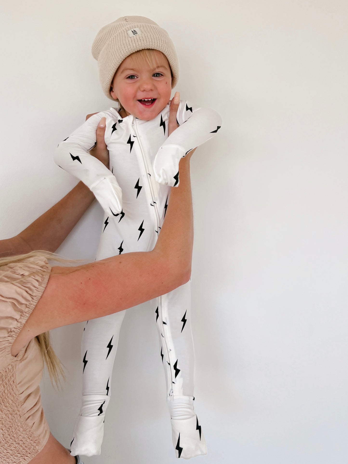 A smiling toddler wearing a Forever French Baby beanie and cozy Bamboo Zip Pajamas in White & Black Lightning Bolt is being held up by an adult's hands against a plain white background. The child looks happy and is looking directly at the camera, radiating joy in their stylish outfit.