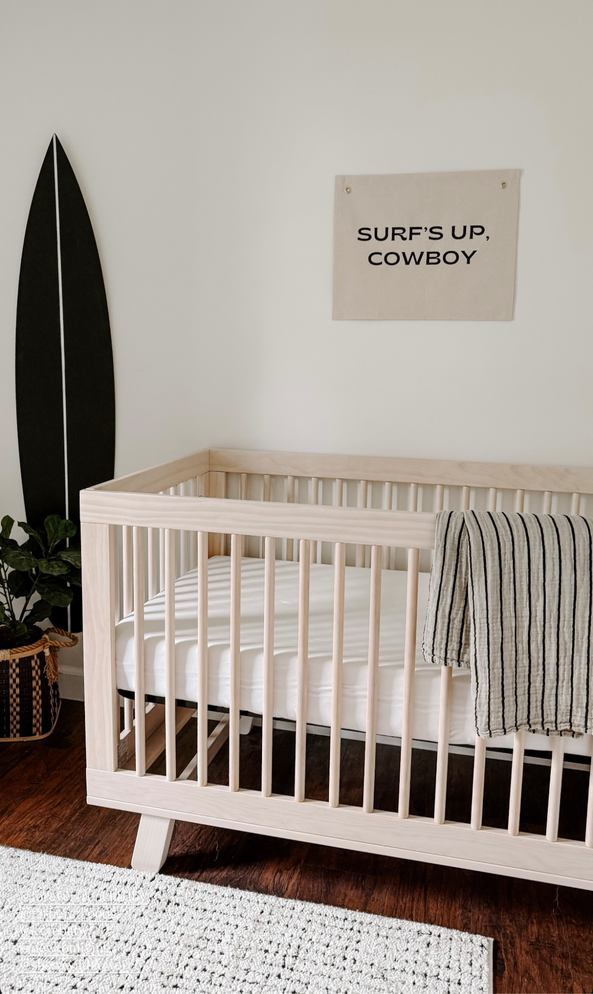 A minimalist nursery features a light wooden crib with the forever french baby Muslin Swaddle | Grey & Black Stripe draped over the side, a dark surfboard against the wall, a potted plant, and a "SURF'S UP, COWBOY" sign.
